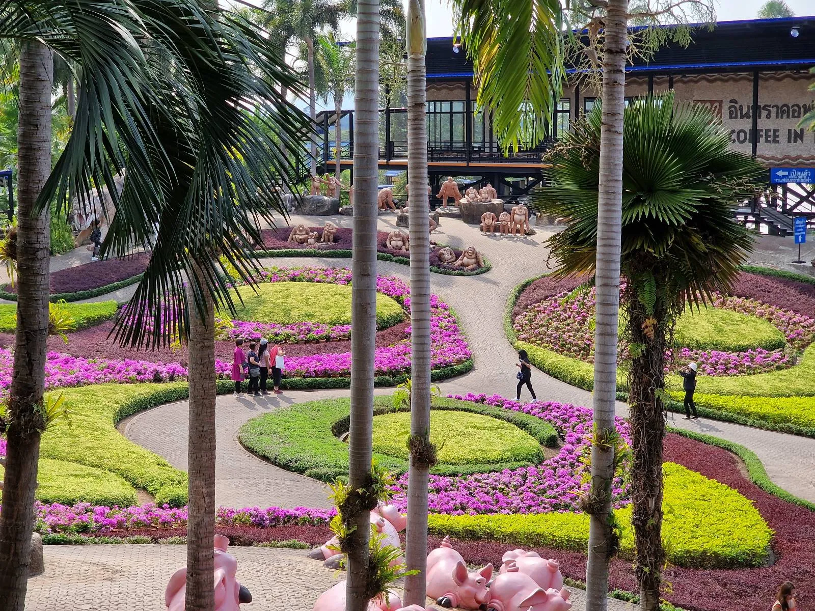A vibrant garden with circular patterns of pink and yellow flowers, surrounded by lush greenery and palm trees. People walk along the curved pathways, and a building is visible in the background.