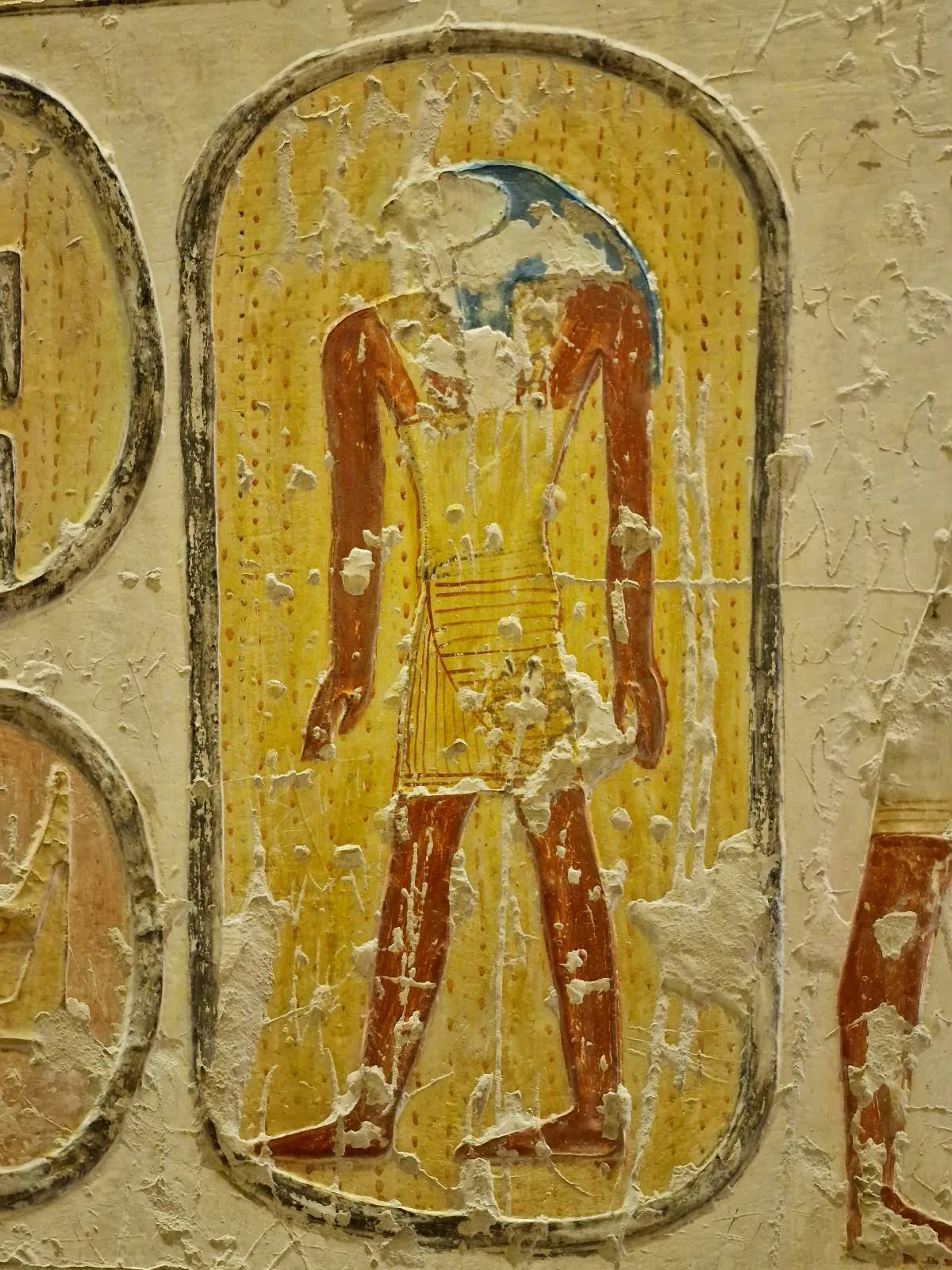 Ancient Egyptian wall painting of a figure with a partially eroded head, wearing a yellow garment. The background contains hieroglyphic text.