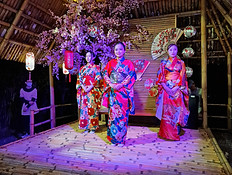 A scenic evening view of a courtyard at Hoi An Memories Theme Park featuring a reflection pool, palm trees, and performers in traditional blue and red attire.