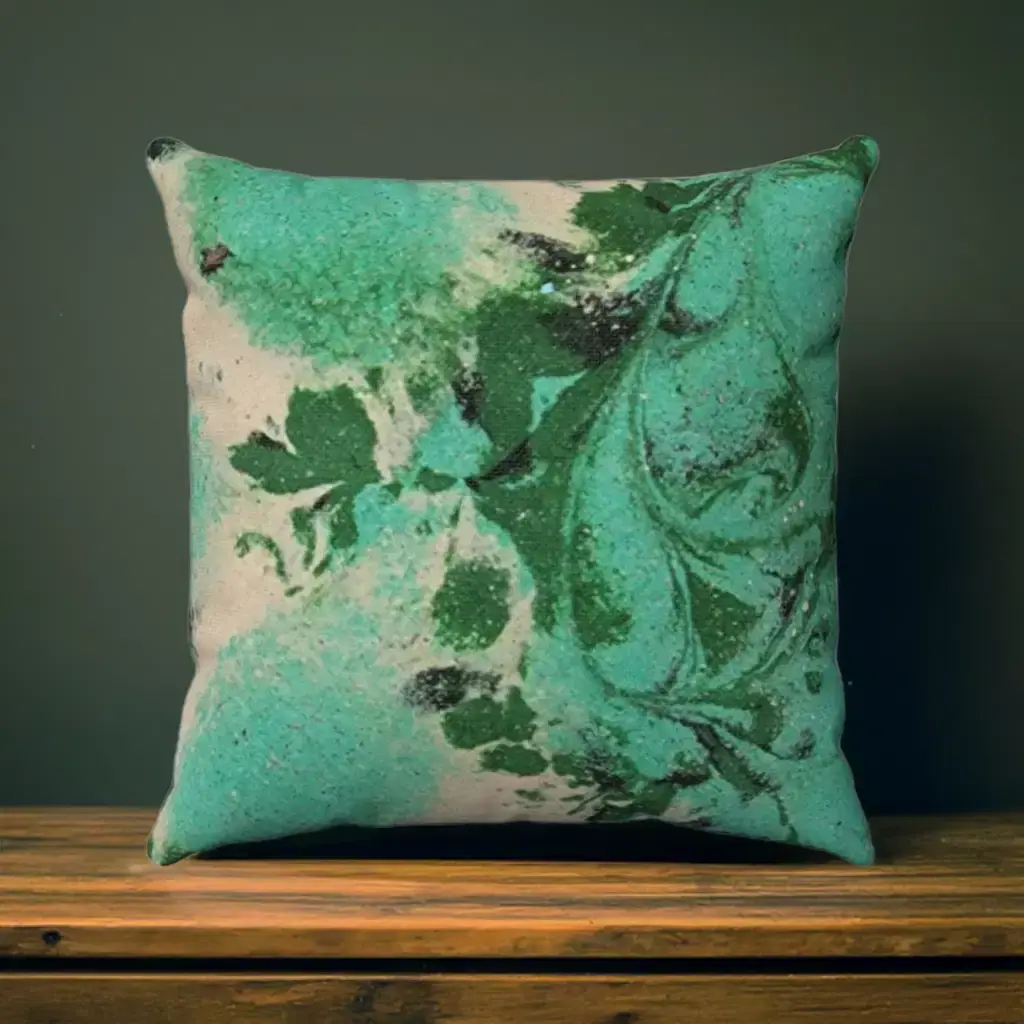 A square cushion with a teal and green abstract floral design rests on a wooden surface against a dark green wall.