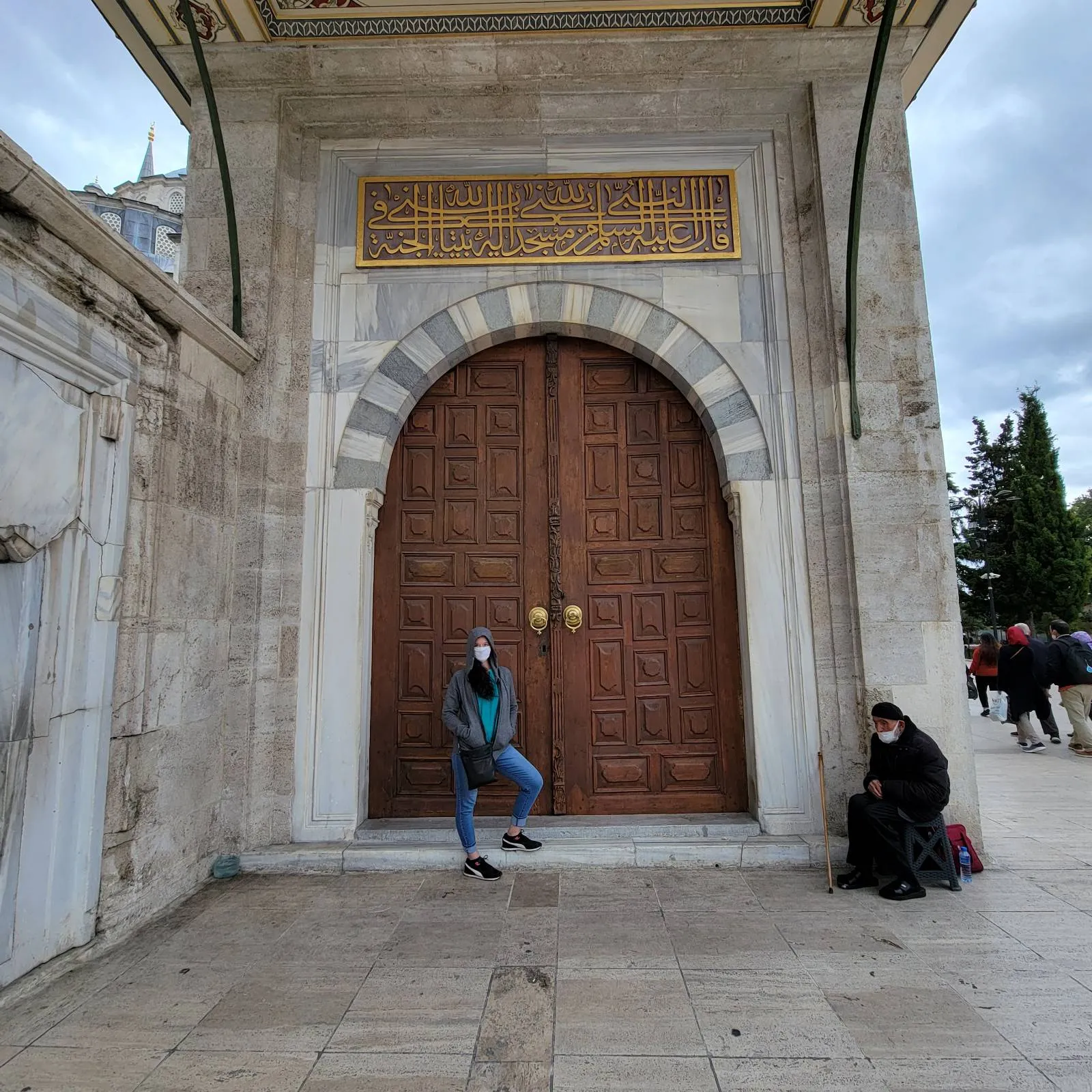 Two people are in front of a large, ornate wooden door with an archway. One stands in the center, while the other sits to the right. The building is made of stone.