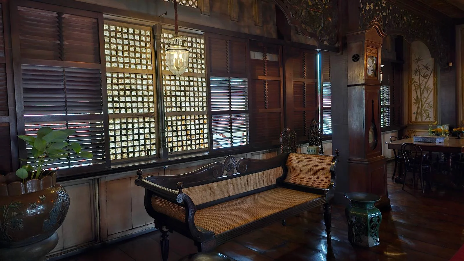 A warmly lit room with wooden floors and traditional wooden furniture. Large windows with intricate lattice designs allow soft light to fill the space. There are decorative plants and a round table with chairs in the background.