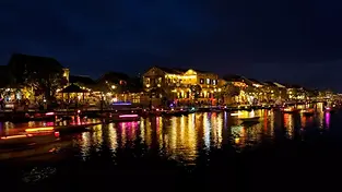 A night view of Hoi An Ancient Town’s waterfront, featuring historic yellow buildings illuminated by warm lanterns reflecting in the Hoai River, with several small boats carrying tourists.