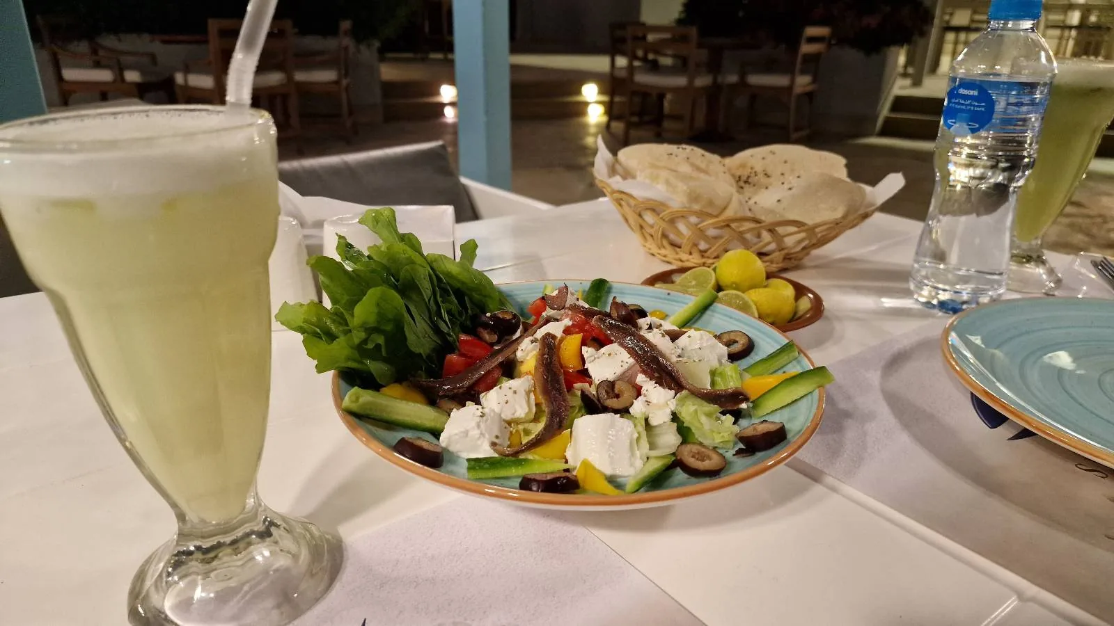 A glass of a light green smoothie next to a plate of fresh salad with cheese, olives, cucumber, and vegetables. In the background, there is a basket of bread and a bottle of water on a white tablecloth.