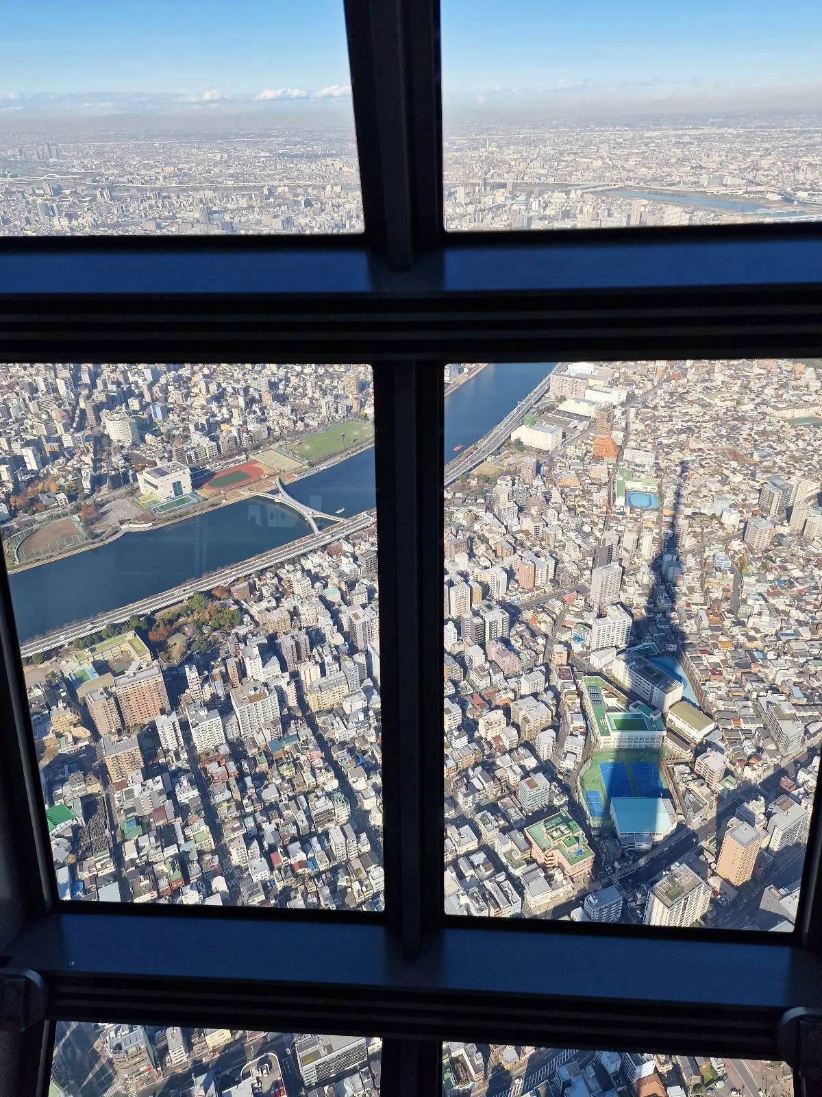 Aerial view of a cityscape seen through a window, with a river and numerous buildings below. The shadow of a tall tower is cast prominently over the urban area. Clear sky above, indicating daylight.
