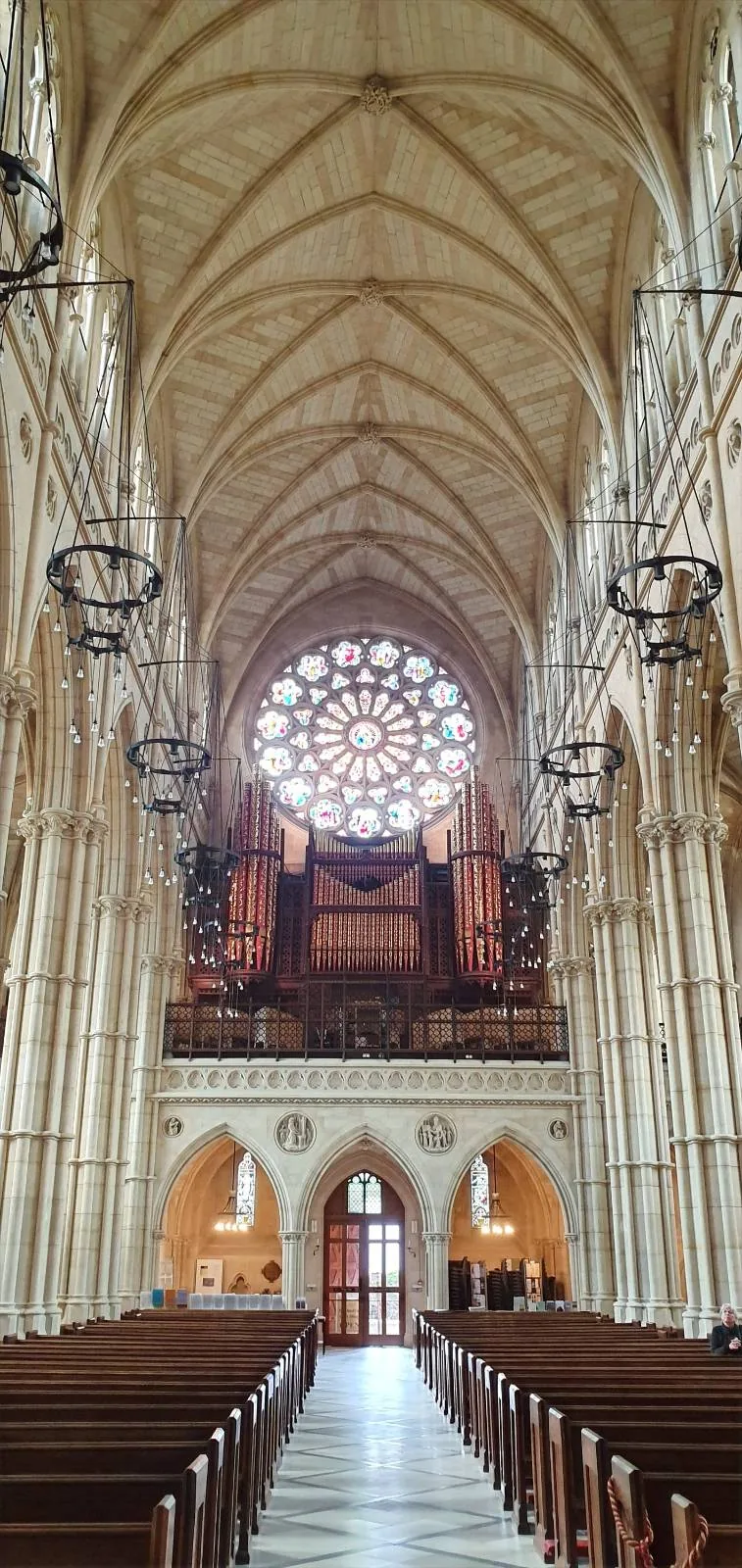 An ornate stone altar with detailed sculptures stands below three stained glass windows depicting colorful religious figures, inside a cathedral. The scene features intricate carvings and a prominent crucifix in the center.
