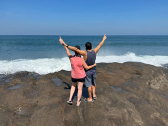 A couple of digital nomads stands on a rocky beach facing the ocean. Both have arms raised enthusiastically, wearing casual summer clothes. Waves crash on the rocks, and the sky is clear and blue, embodying the spirit of Closer Lives with their adventurous journey.