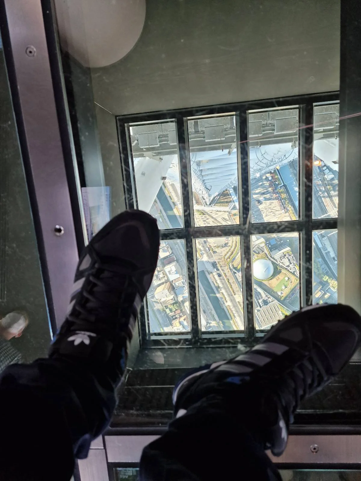View through a glass floor showing a cityscape from above. Two feet in sneakers are visible, standing on the transparent surface, with buildings and streets far below.