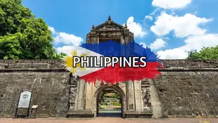 Entrance gate of an old stone fortress with intricate carvings and coat of arms, surrounded by greenery; the word "PHILIPPINES" overlays a painted Philippine flag at the bottom center.