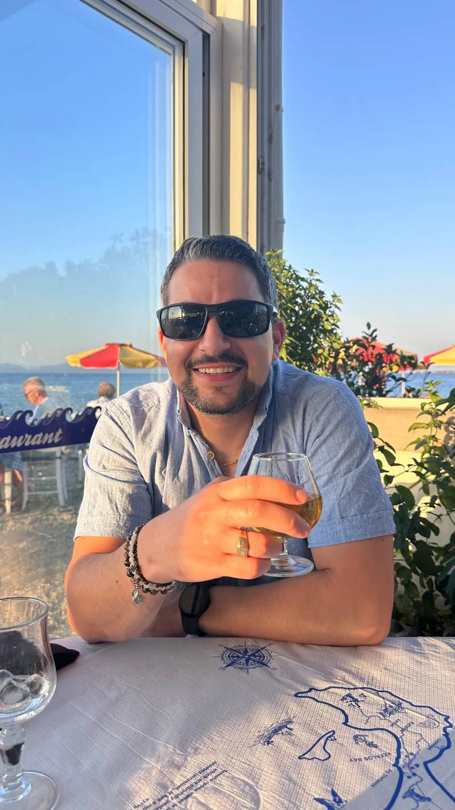 A man wearing sunglasses and a short-sleeved shirt sits at an outdoor table, smiling and holding a drink. The setting is sunny, with a view of the sea, red umbrellas, and greenery in the background.