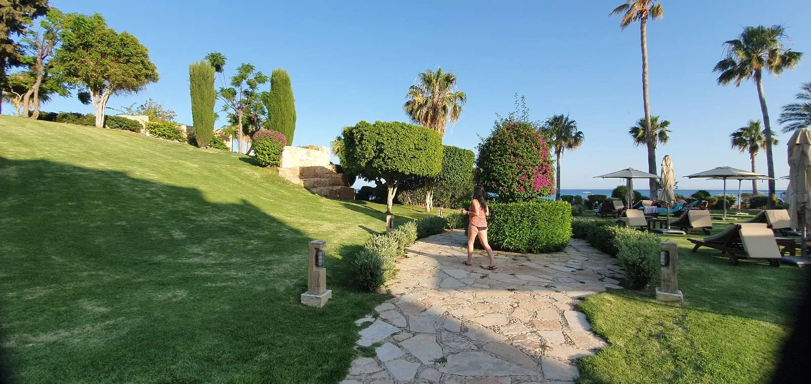 A person walks along a stone path in a lush garden with neatly trimmed bushes, palm trees, and a grassy hill. In the background, there are white umbrellas and lawn chairs near the sea under a clear blue sky.