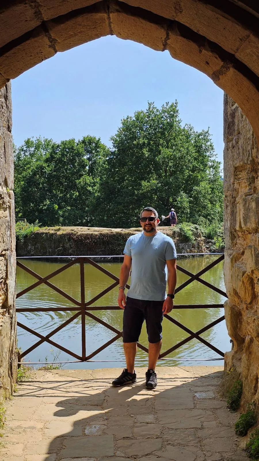 A man in a light blue shirt and black shorts stands under a stone archway by a metal gate, with a calm river and lush green trees in the background on a sunny day.