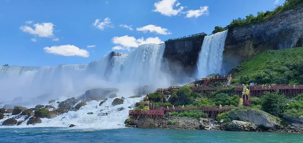 Massive waterfall with tourists in yellow ponchos on red wooden walkways. Blue sky with clouds, lush greenery, and flowing water below.