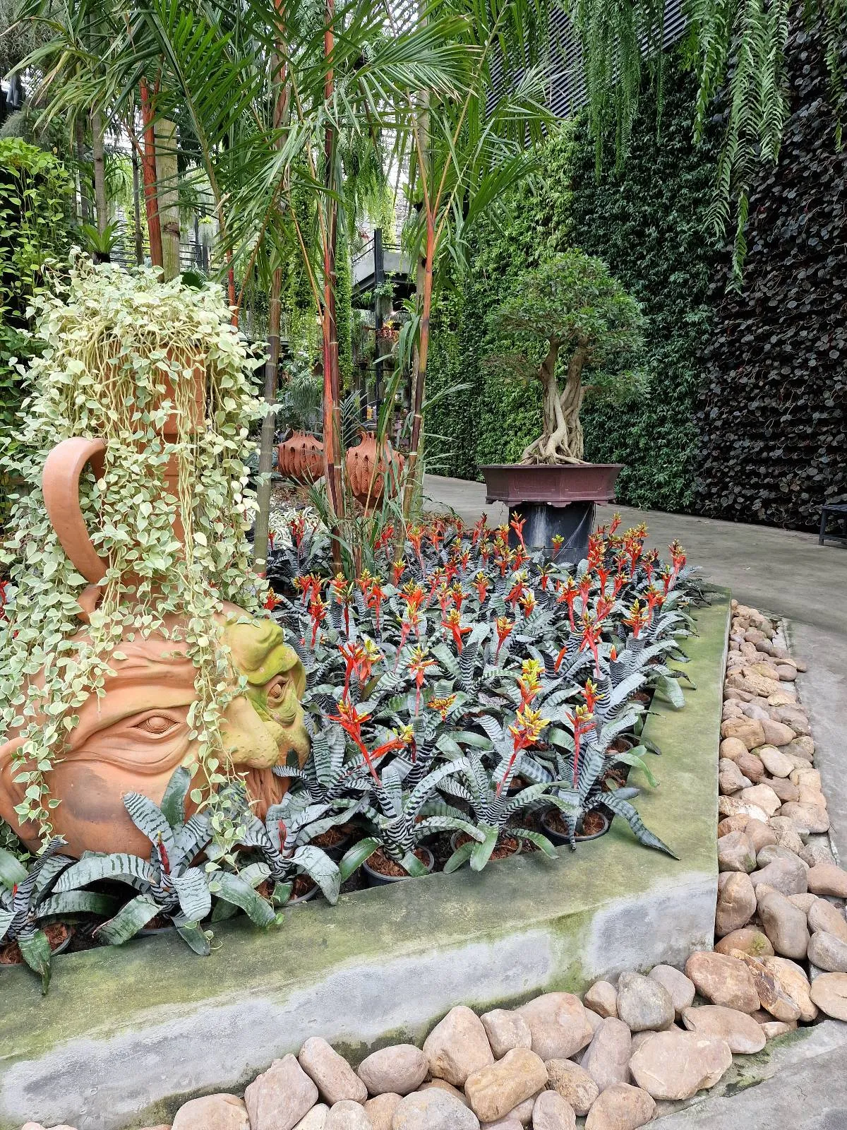A garden scene with a large decorative pot resembling a face, overflowing with cascading greenery. Bright red flowers and leafy plants surround the pot. A small bonsai tree is placed on a stand nearby, with a backdrop of a lush vertical garden.