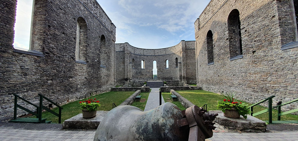 Stone ruins with tall walls and arched windows under a blue sky. A large bell is in the foreground, surrounded by red flowers.