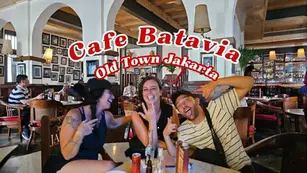 Three people laughing at a table in Cafe Batavia, Old Town Jakarta. Cozy interior with warm lighting and framed photos. Cafe name in bold text.