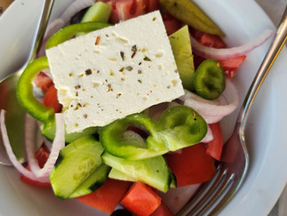 A bowl of Greek salad with chopped cucumbers, tomatoes, green bell pepper, red onion, and a slice of feta cheese on top, garnished with herbs. A fork and spoon rest on the edge of the bowl.