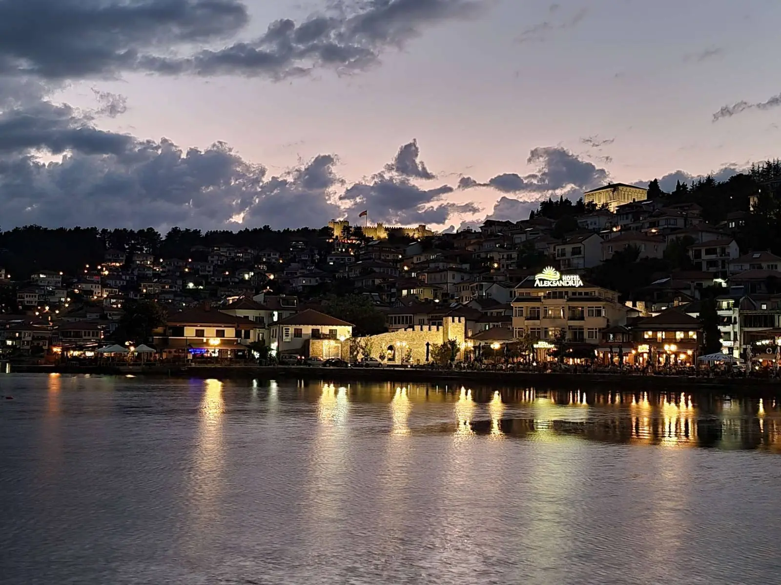 Evening view of a coastal town with buildings lit up along the water. The sky is a mix of dusk colors with scattered clouds. Reflections of the lights shimmer on the calm water's surface. Hills are visible in the background.