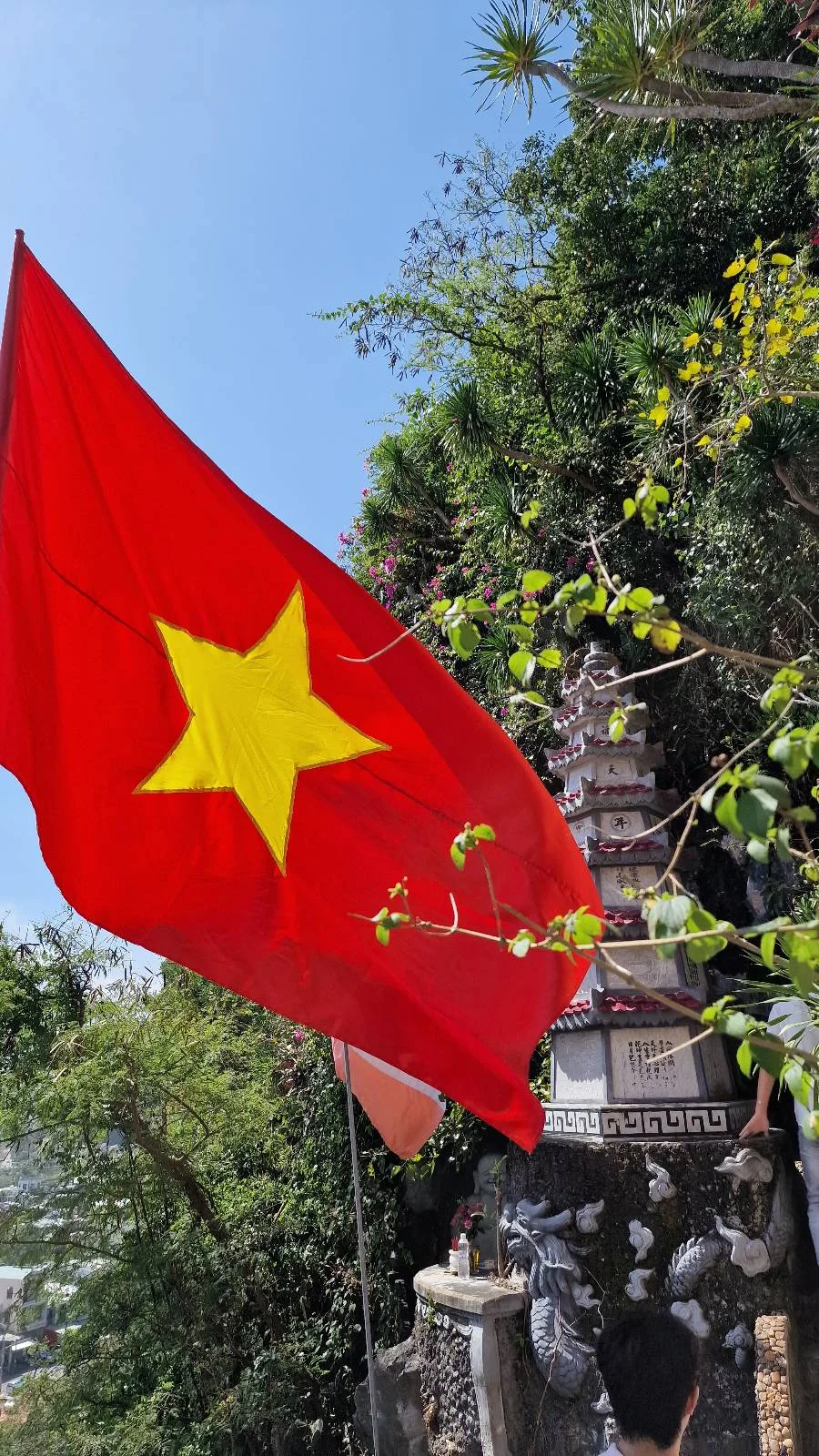 A red flag with a yellow star flies near a monument surrounded by greenery and plants under a clear blue sky.