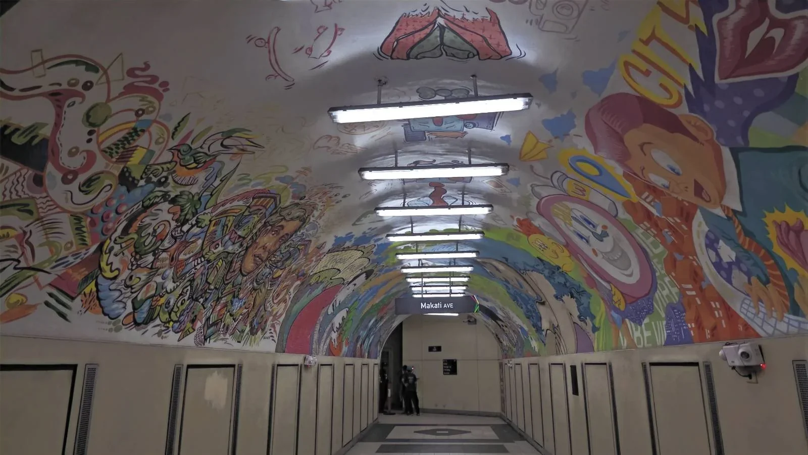 A hallway with a curved ceiling covered in colorful murals and fluorescent lights, with doorways lining both sides and a person standing at the far end.