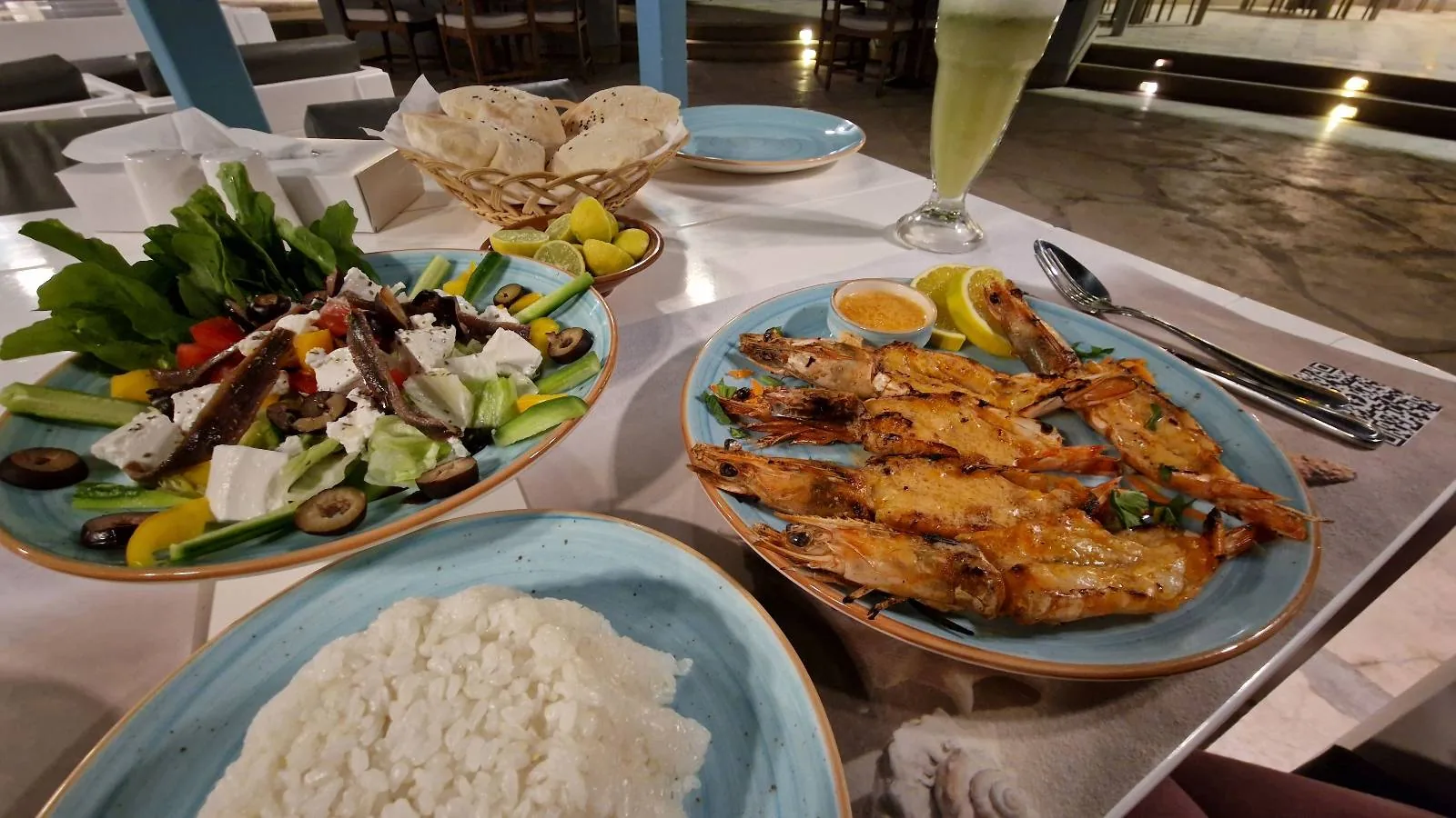 Plates of grilled shrimp, a mixed vegetable salad, white rice, and a glass of juice are set on a dining table.