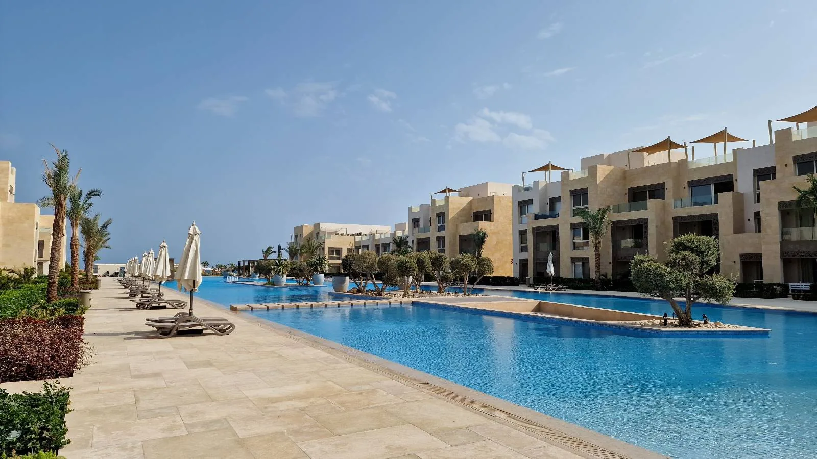 A large swimming pool stretches between modern, beige-colored buildings under a blue sky. Lounge chairs and umbrellas line the poolside, and a few palm trees are scattered around the area.