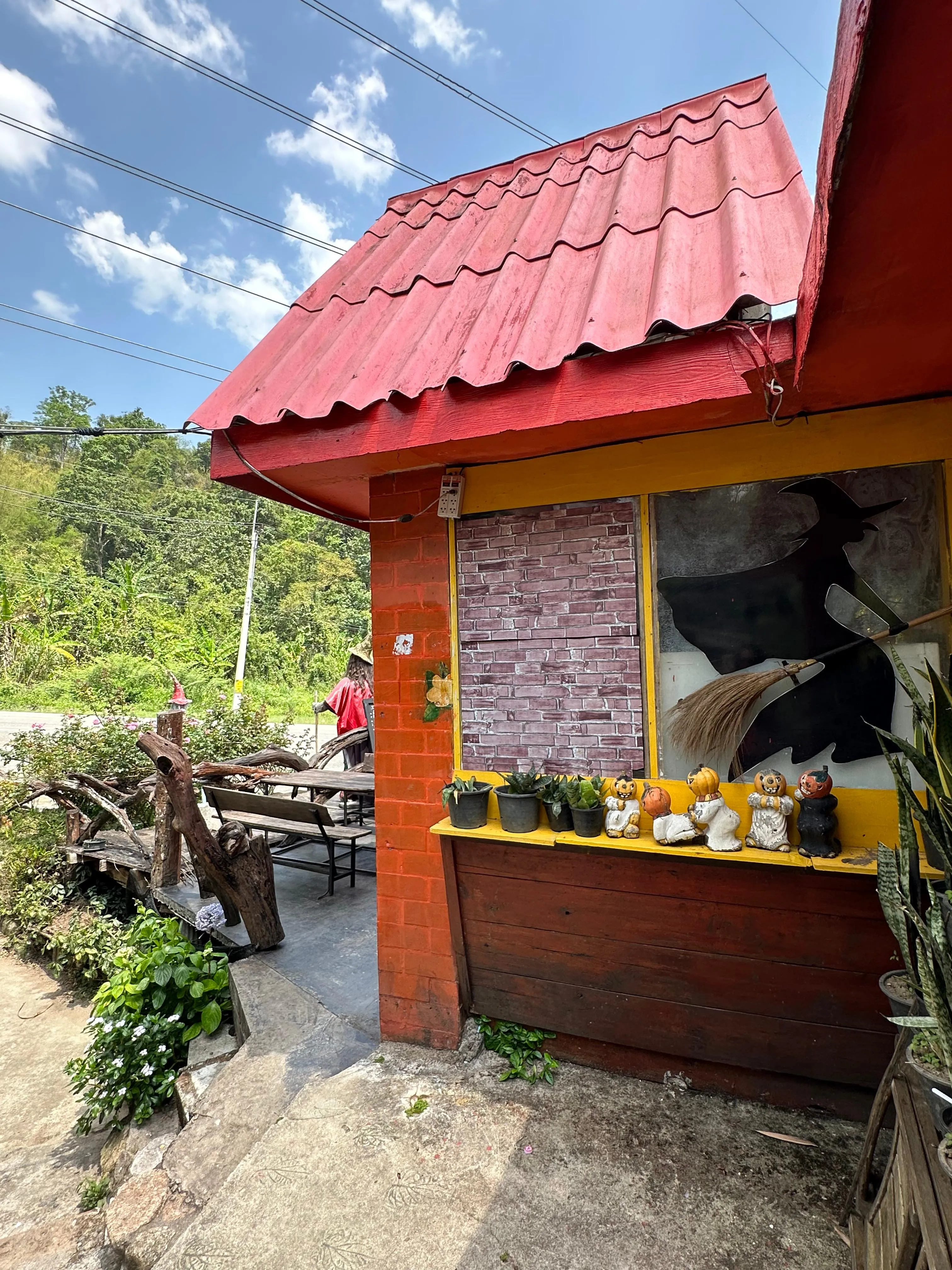 A small roadside stand with a red roof displays fruits and jars on the window ledge. Outdoor seating with tables and chairs is visible to the side, surrounded by greenery under a blue sky with scattered clouds.