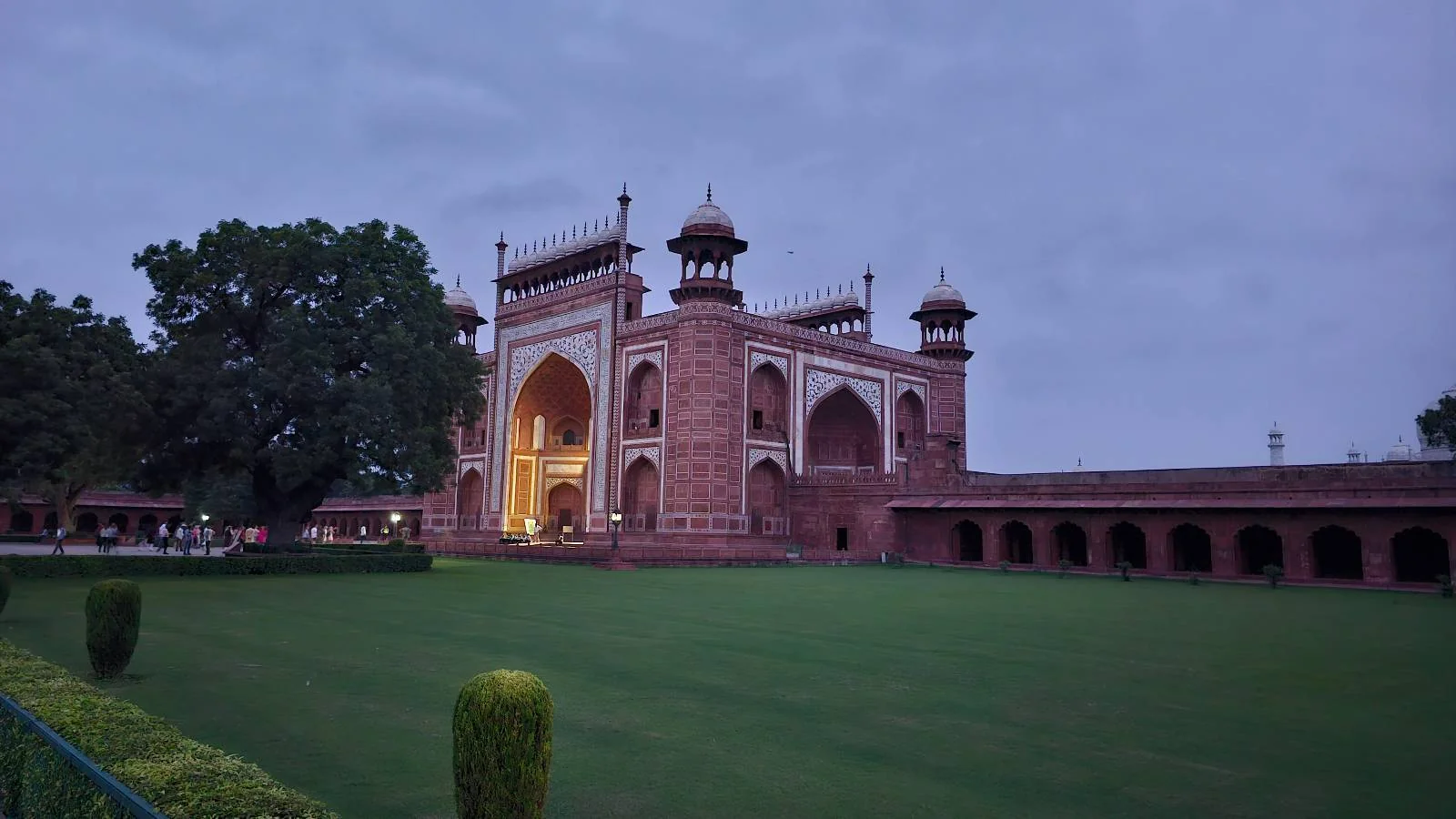 The image shows a large red sandstone gateway with domes and arches, set against a cloudy sky, surrounded by a manicured lawn and trees, with people walking near the entrance.