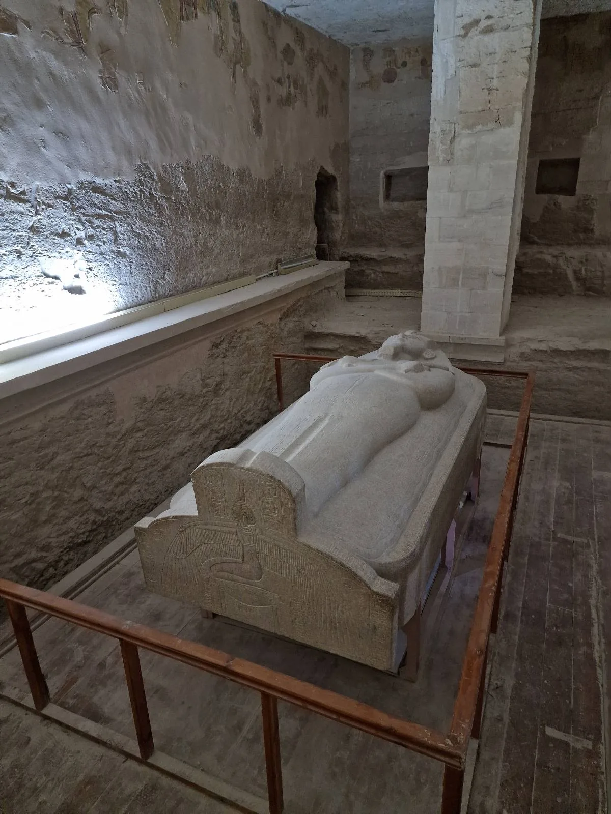 A stone sarcophagus is displayed within a fenced area in a dimly lit, ancient, and worn stone room.