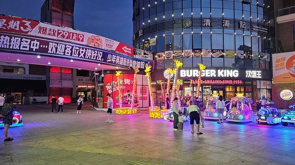 People in a plaza with neon giraffe-themed rides. A Burger King with bright signage is in the background. Evening setting with vibrant lights.