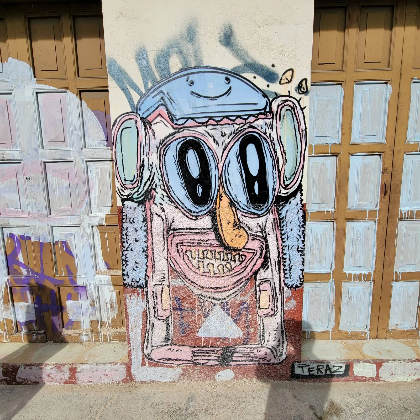 A colorful graffiti mural on a wall depicts a cartoonish face with large eyes, a long nose, and a wide grin. The design includes elements resembling headphones and a hat. The background has wooden doors with faded paint.