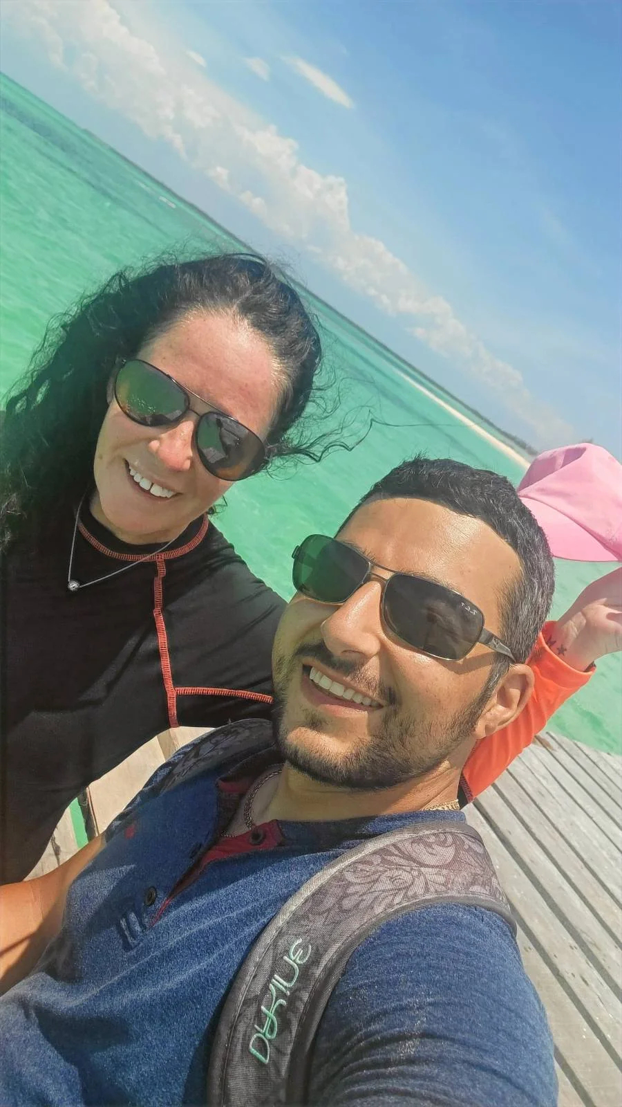 Two people wearing sunglasses smile at the camera while standing on a wooden pier. They are near an ocean with clear turquoise water under a blue sky. One person has a backpack on their shoulder.