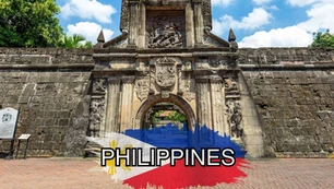 Entrance gate of an old stone fortress with intricate carvings and coat of arms, surrounded by greenery; the word "PHILIPPINES" overlays a painted Philippine flag at the bottom center.