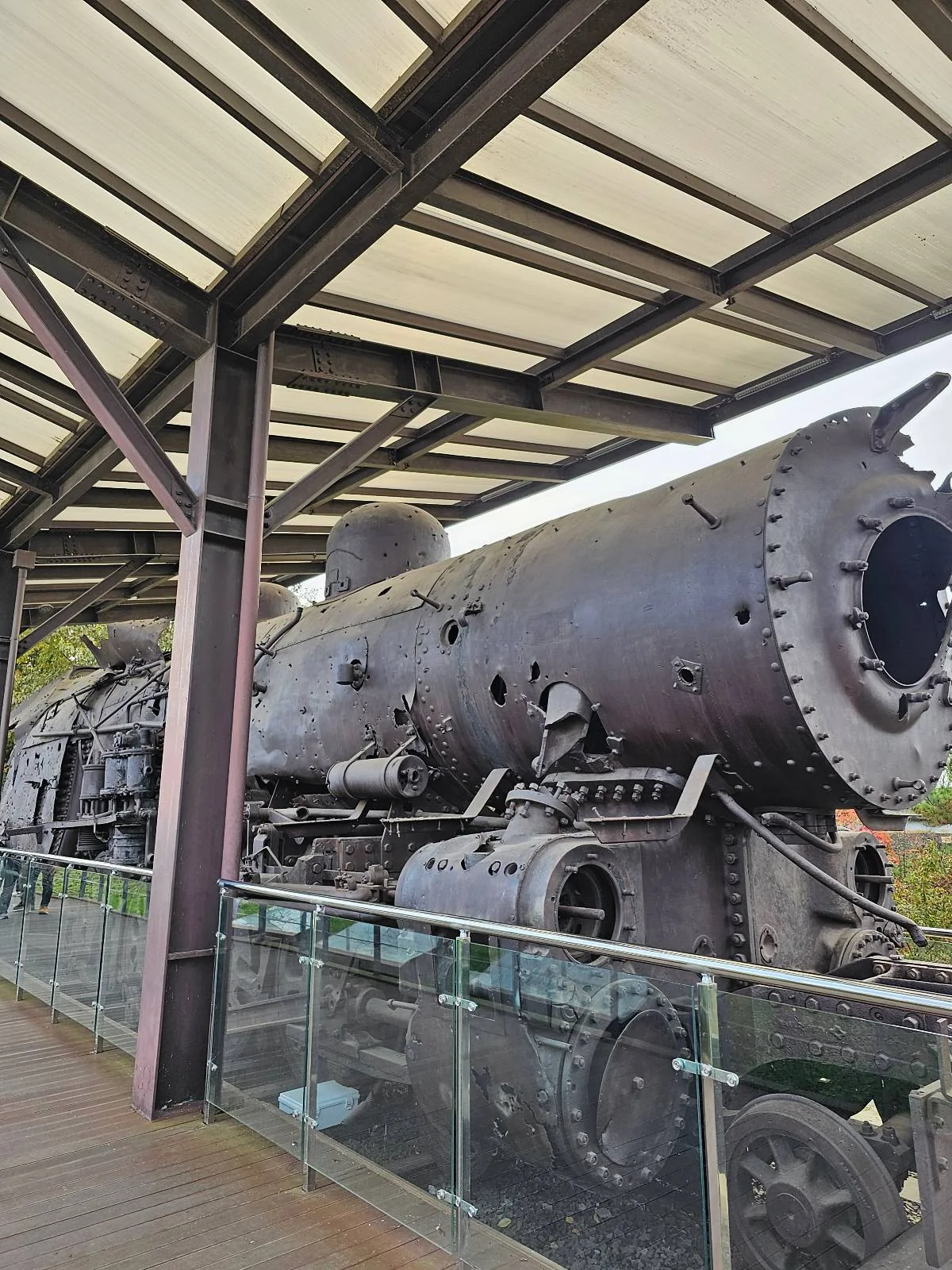 A large, rusted steam locomotive displayed under a covered structure. The engine's stripped exterior reveals mechanical components. Glass barriers surround it, and the roof provides partial shelter.