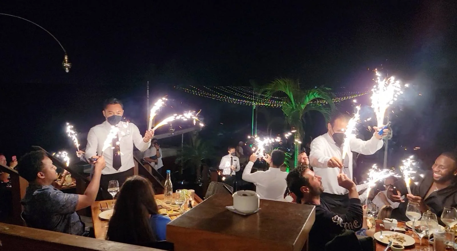 People are dining outdoors at night. Several waiters hold sparkling fireworks, creating a festive atmosphere. There are tables with food and drinks, and diners are engaging with each other in a lively setting.