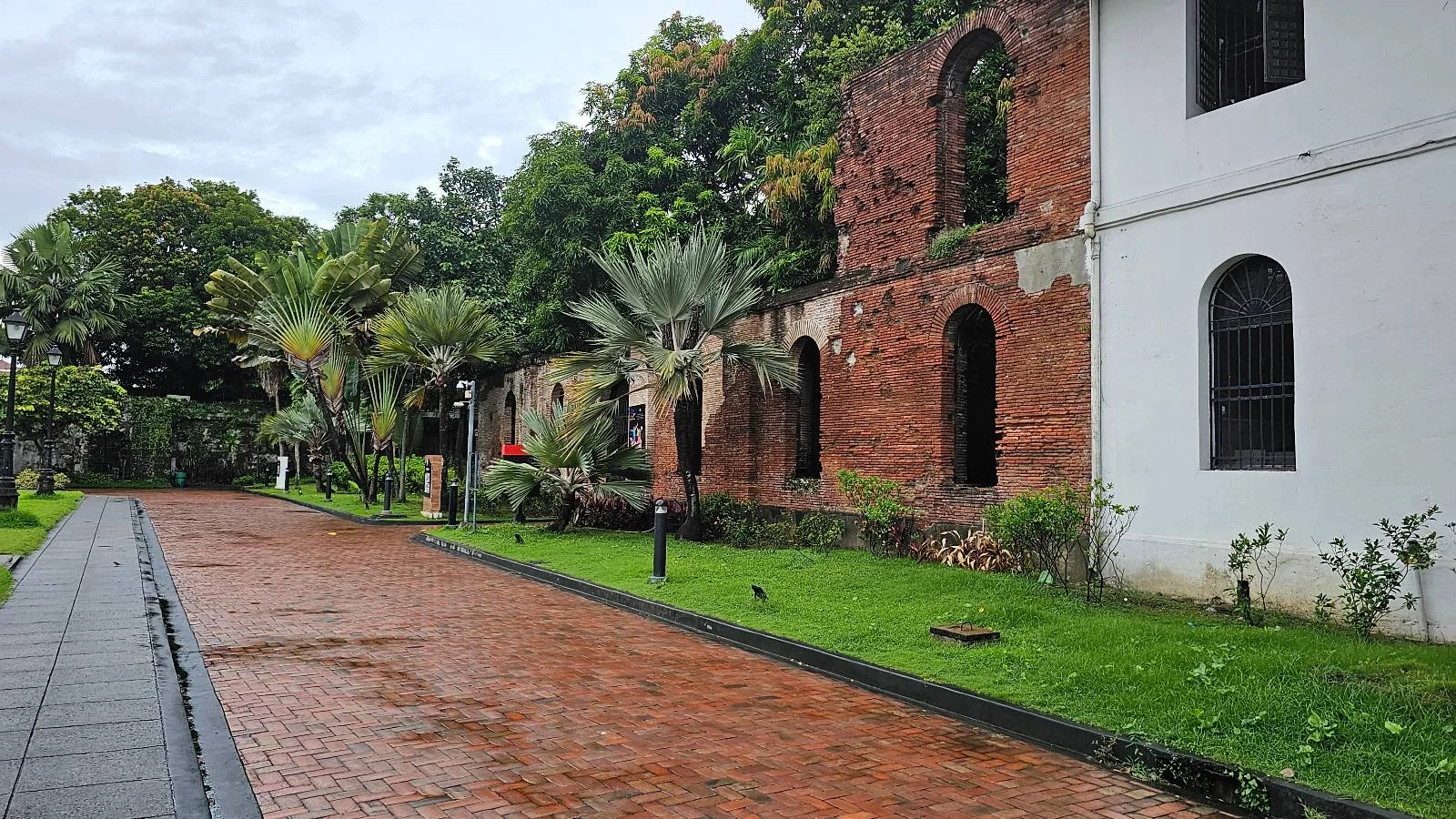 A brick pathway runs alongside old red-brick ruins and a white building, surrounded by green grass, palm trees, and lush vegetation under a cloudy sky.