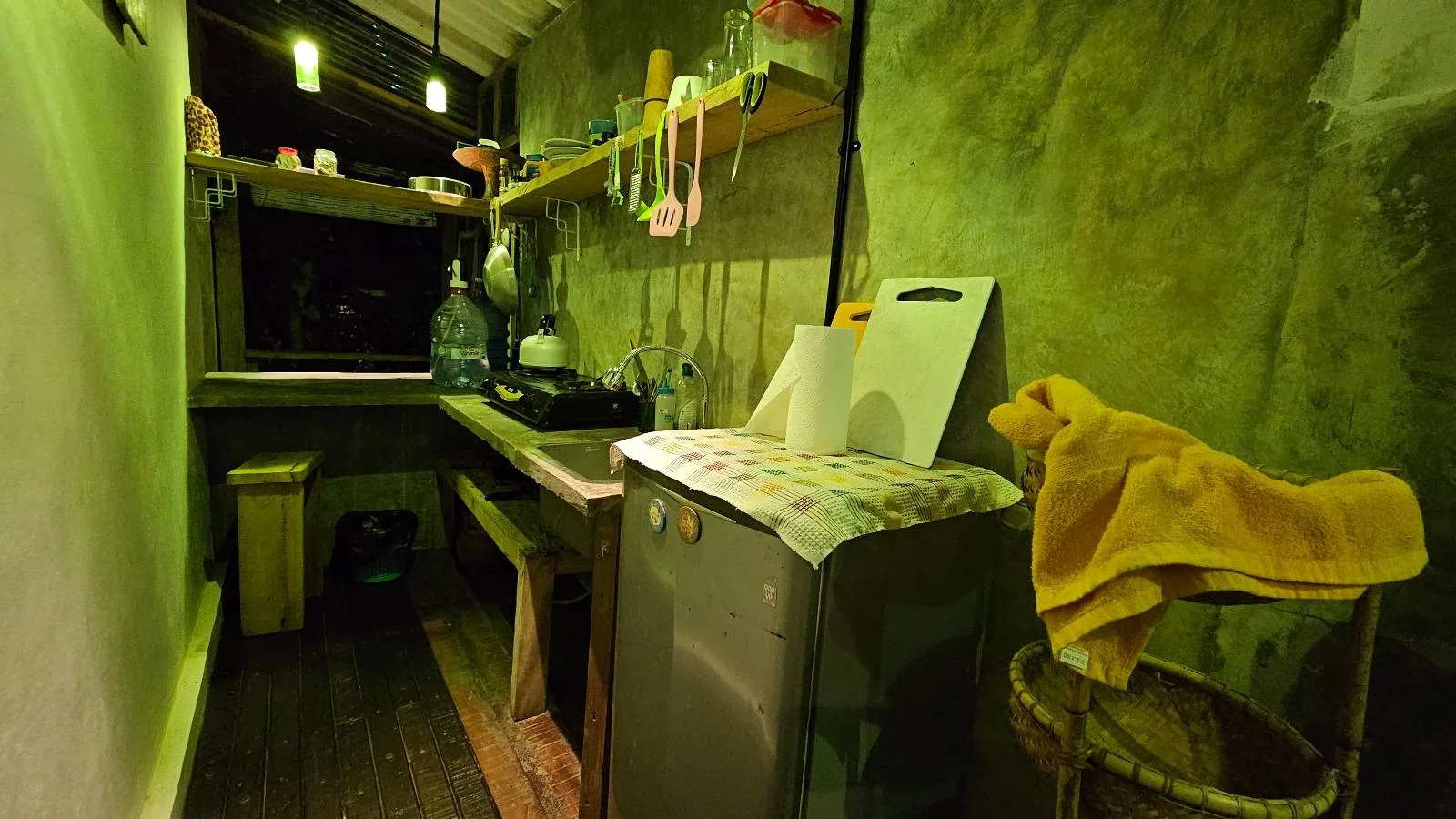 A narrow, dimly lit kitchen with green walls, a small counter, and open shelves holding kitchen items. There is a mini fridge, a basket with a yellow towel, and various utensils hanging above the sink area.