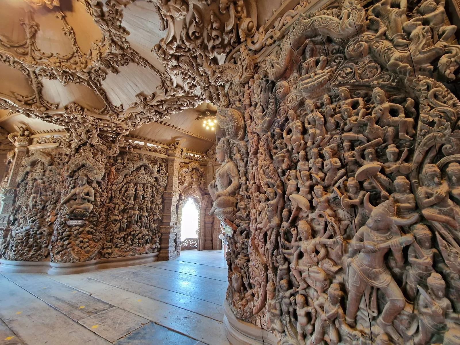 Intricately carved wooden pillars and walls inside a temple, featuring detailed reliefs of people, animals, and elaborate patterns, with natural light streaming through an arched doorway in the background.