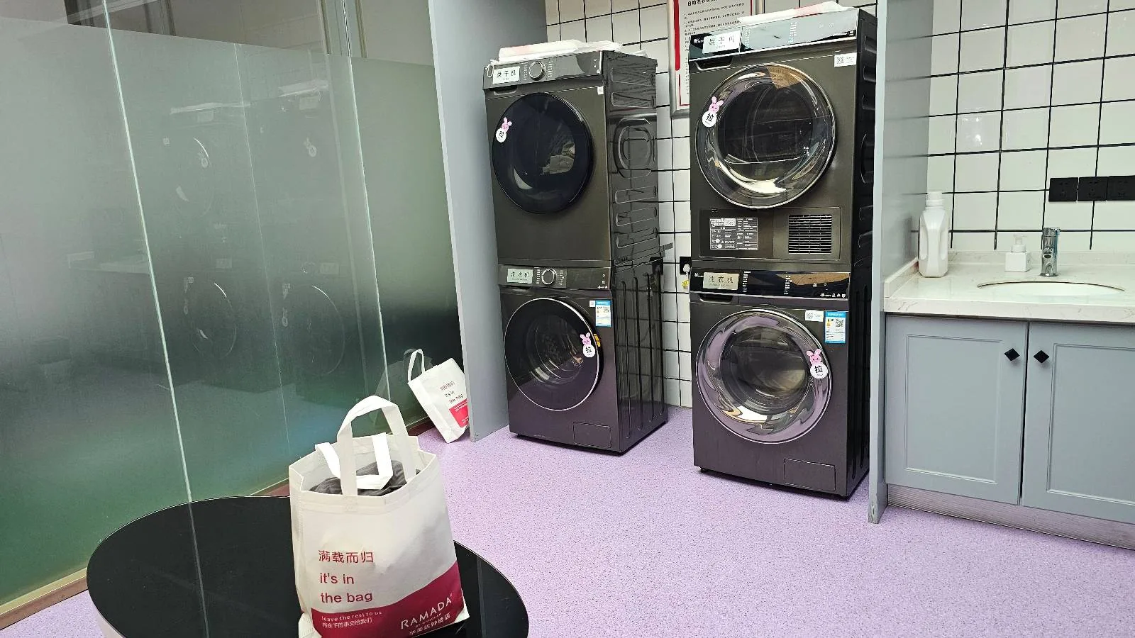 A modern laundry room with two stacked washing machines and dryers against a tiled wall, a sink with cabinets, and a table with a shopping bag in the foreground.