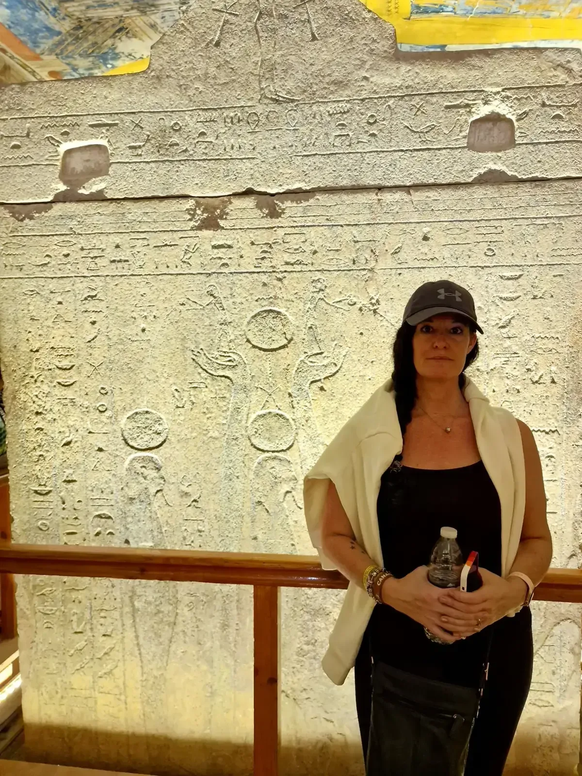 A person wearing a dark baseball cap and white shawl stands in front of a large ancient stone tablet with carvings. They hold a small item in their hands. The scene is indoors with warm lighting.