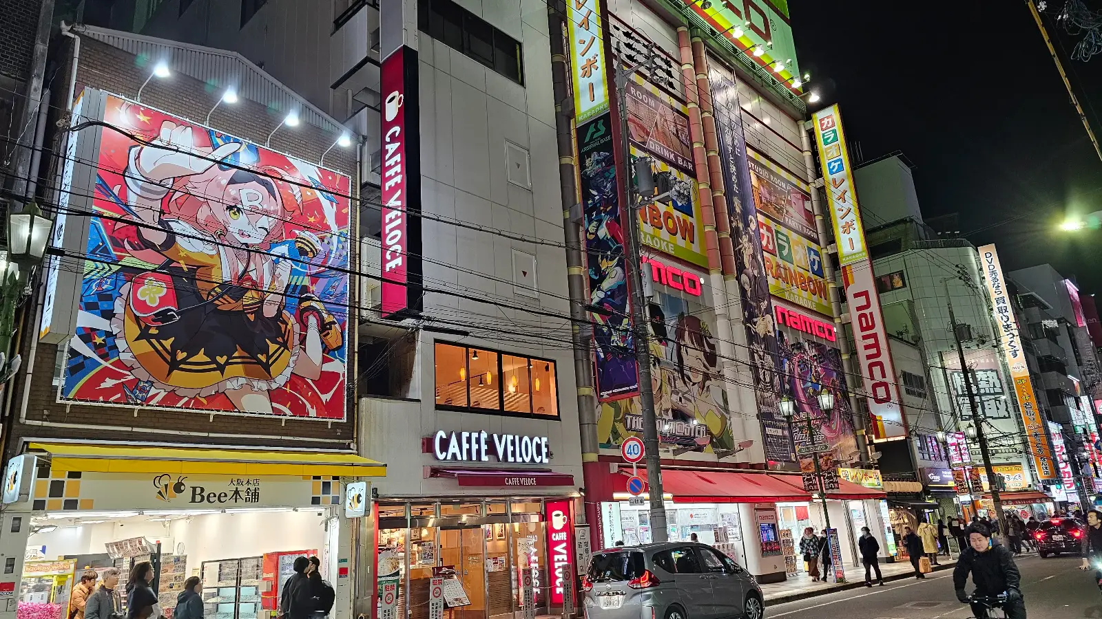 Colorful street scene at night in Akihabara, Tokyo, with bright anime billboards, illuminated shop signs, and people walking on the sidewalk in front of various stores and cafés.