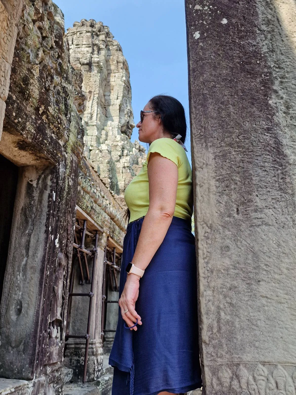 A woman in a yellow top and blue skirt stands sideways next to ancient stone ruins, looking toward the left, with weathered walls and a clear blue sky in the background.