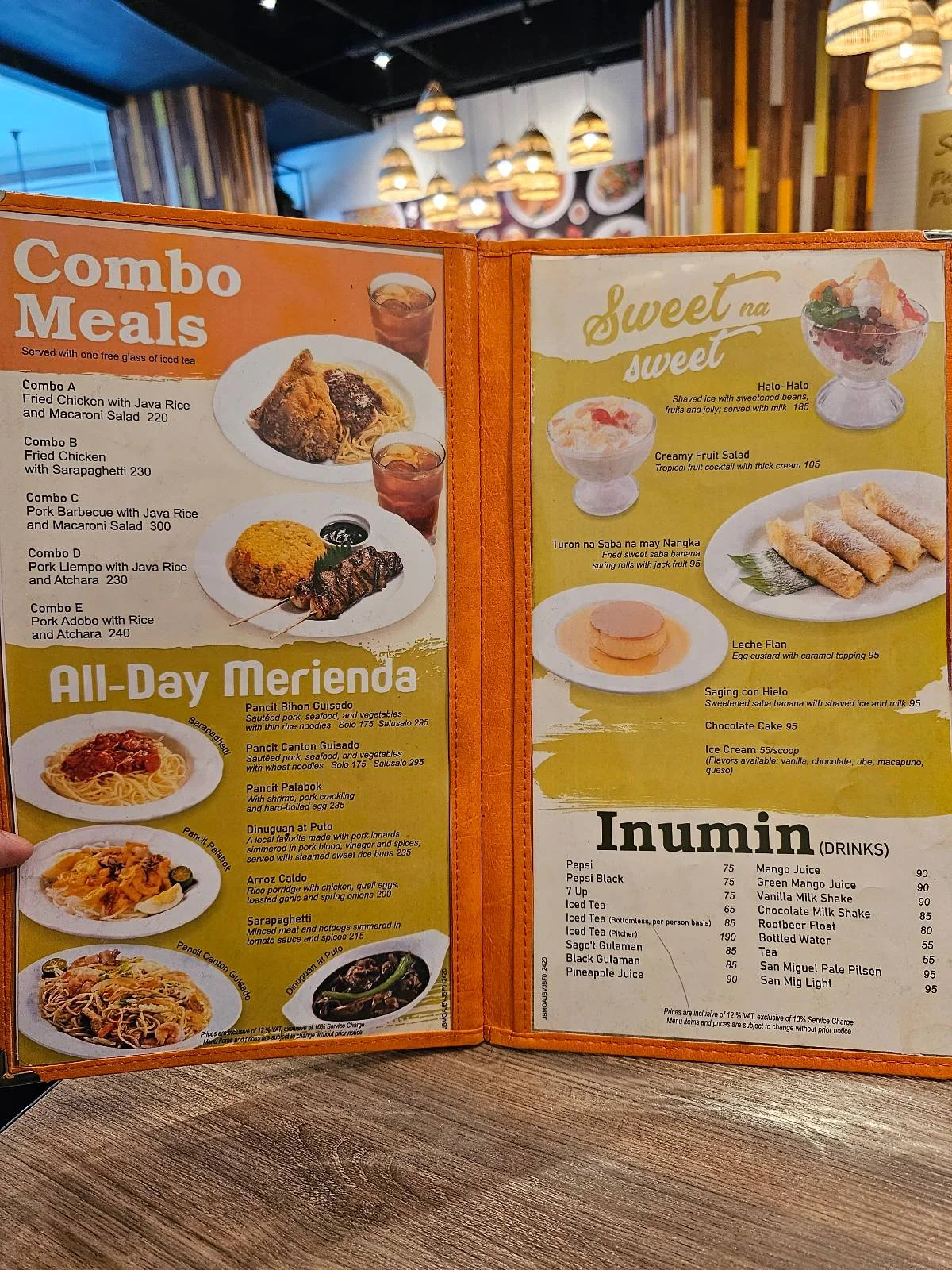 A restaurant menu displayed on a wooden table offers combo meals, all-day snacks, desserts, and drinks. The menu shows various dish images, including rice meals, pastries, and beverages, in a colorful design.