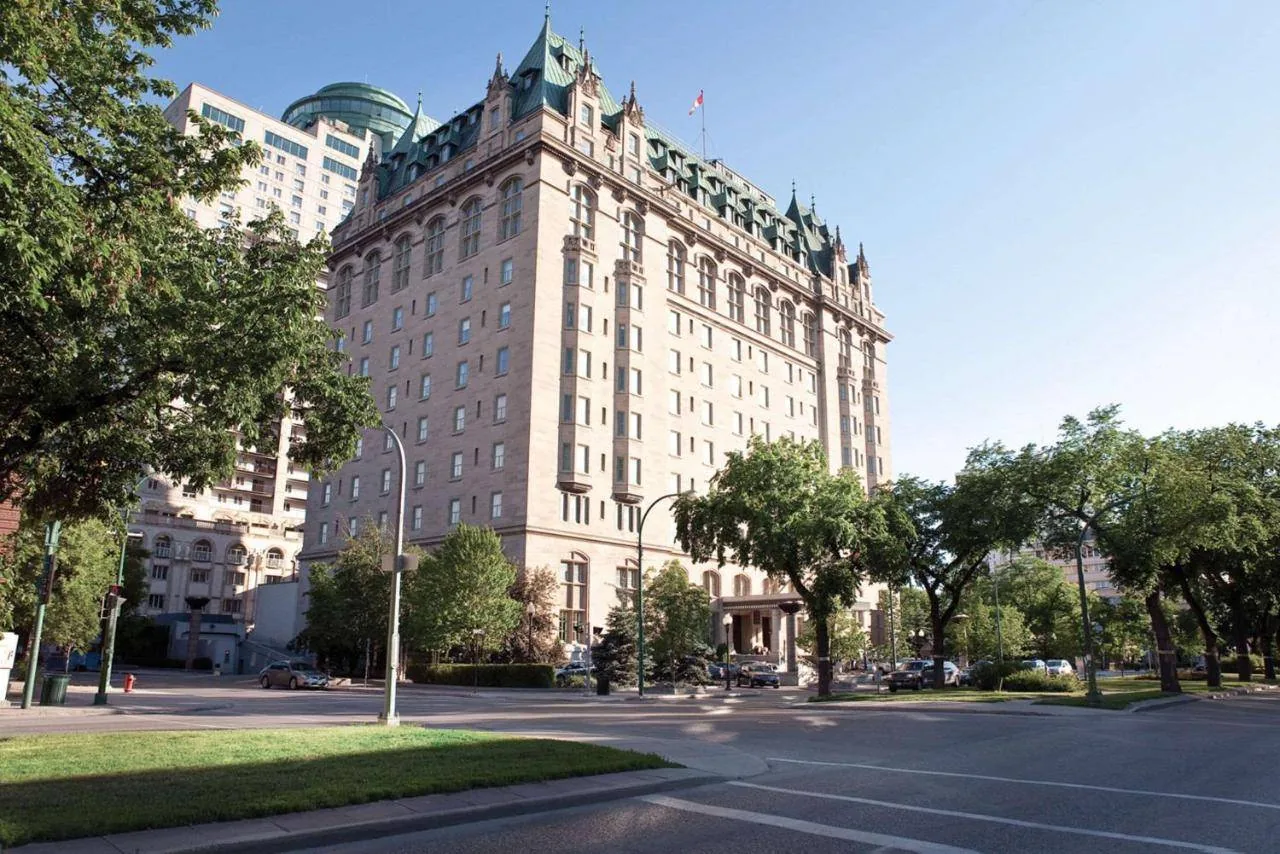 A grand, multi-story historic hotel with a chateau-like design stands on a street corner. It's surrounded by trees and adjacent to a modern building under a clear blue sky.