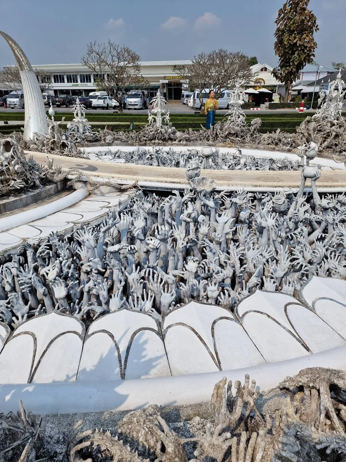 A detailed sculpture with intricate white carvings and spiky structures outdoors. In the background, cars are parked near a building, and a person in a yellow top and blue jeans is walking. The sky is clear and sunny.