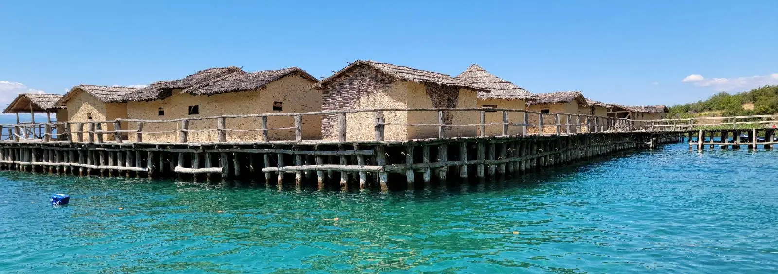 A series of overwater bungalows with thatched roofs extend over clear blue water under a bright blue sky. The bungalows are connected by wooden walkways. A small buoy floats in the water nearby.