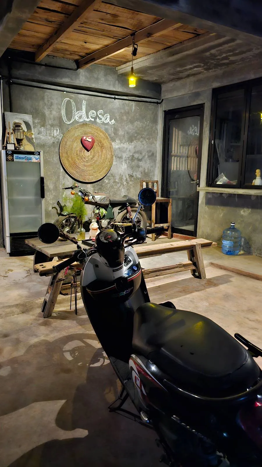 A dimly lit home with a motorcycle parked indoors. The wall features a large straw decoration with "Odesa" and a red heart. A wooden picnic table holds plants and decor. Industrial gray walls and lighting create a cozy ambiance.