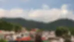 Rooftops of a colorful town with hills and lush greenery in the background under a cloudy sky. The scene is calm and scenic.