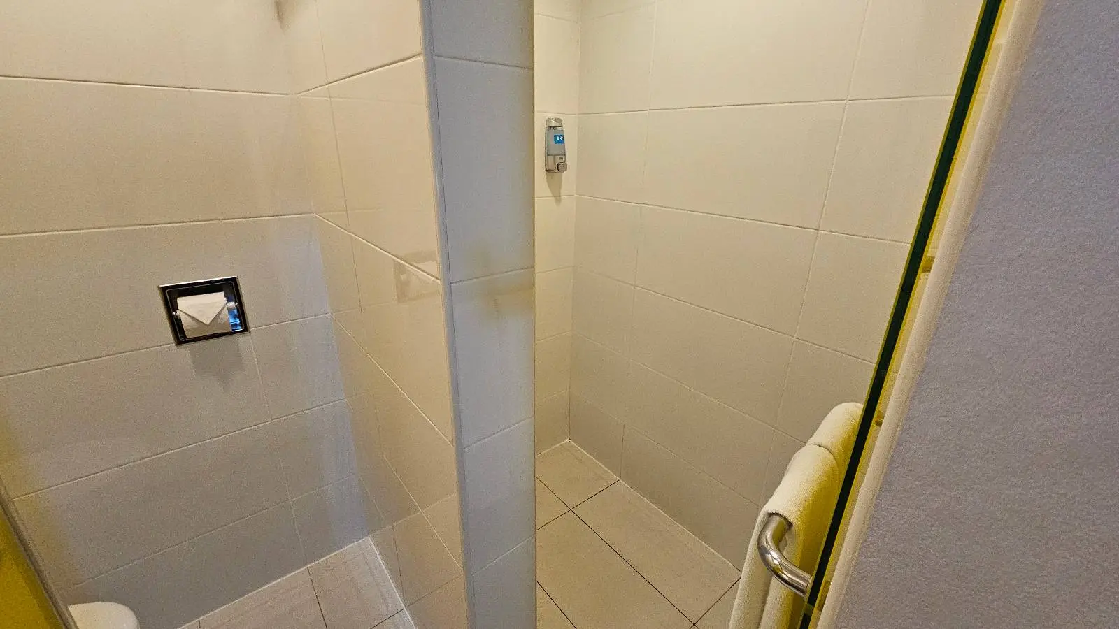 A small, tiled bathroom with a walk-in shower, a mounted soap dispenser, a towel hanging on a rack, and a toilet paper holder attached to the wall.