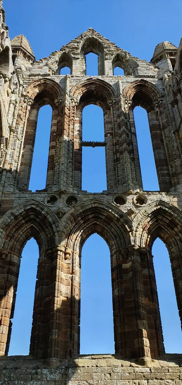 A view of tall, arched stone windows in a historic, Gothic-style building against a clear blue sky. The structure is weathered, highlighting its age and architectural design.